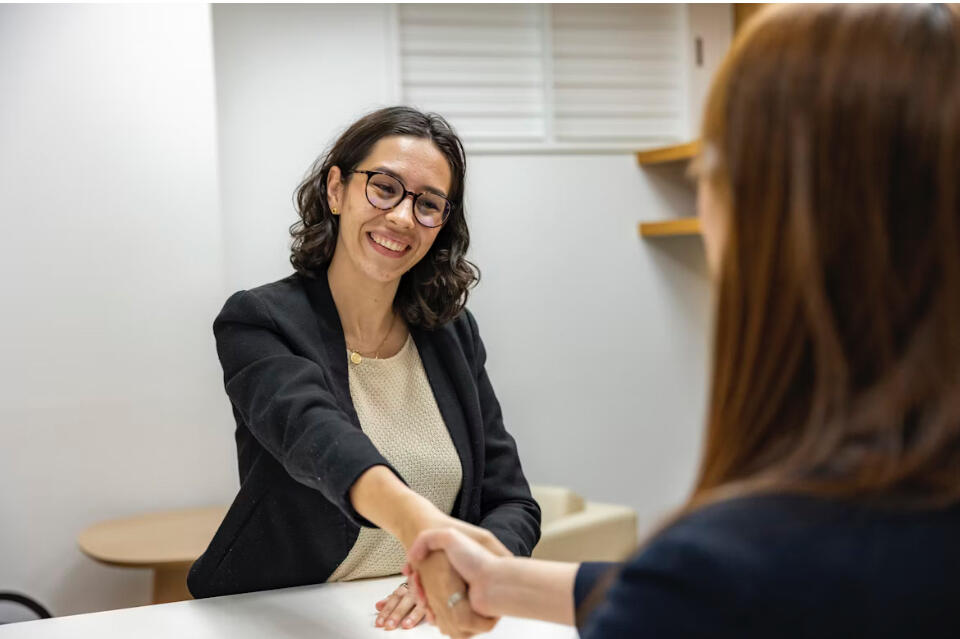 Image of a special education advocate with dark hair, glasses, and wearing a black suit smiling broadly while shaking hands with a younger woman with long dark hair across the advocate’s office desk.