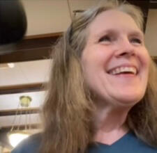 Casual photo of Refine Disabilities founder Donna Earnest. She is a white woman in her late 60’s, with shoulder length brown mixed with gray hair, wearing light makeup. She is smiling with joy at something off camera. She is indoors, wearing a dark teal te